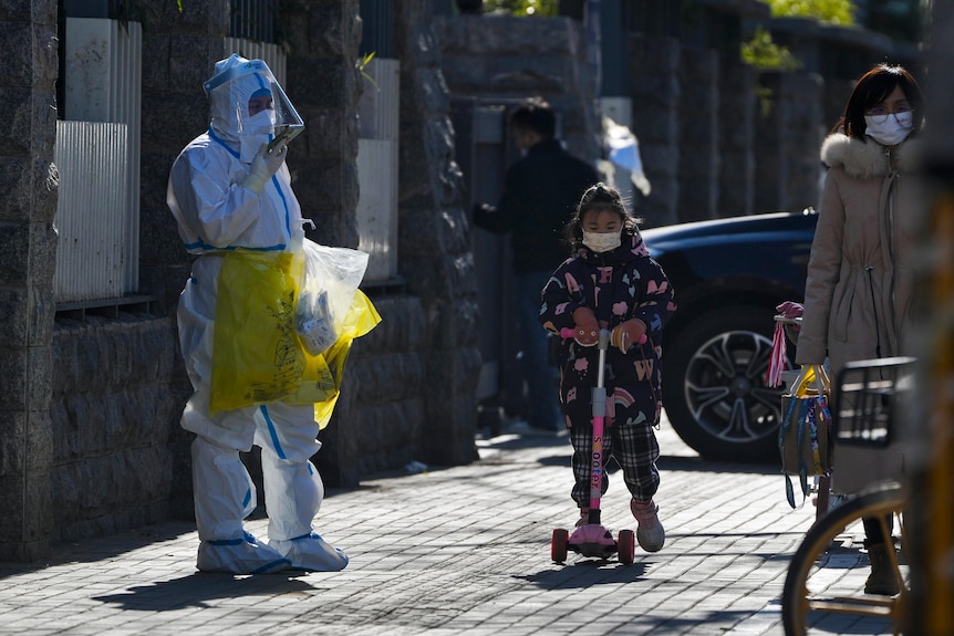 A Chinese child wearing a face mask and riding on a scooter passes a man in protective clothing holding yellow biowaste bags.