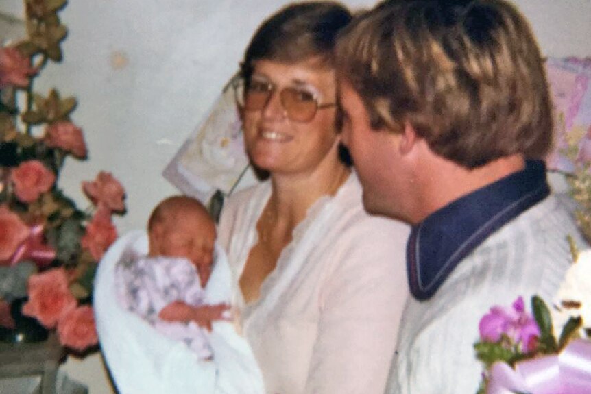 Lyn Dawson sits on bed holding a baby, back of Chris Dawson's head can be seen