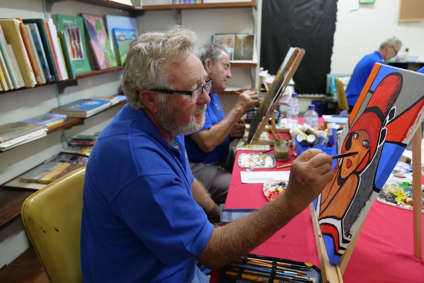 A man sits at a table painting on a canvas.