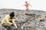 A man squatting on a rocky bank and placing his hand in a stream of water while a woman jumps over it, white background