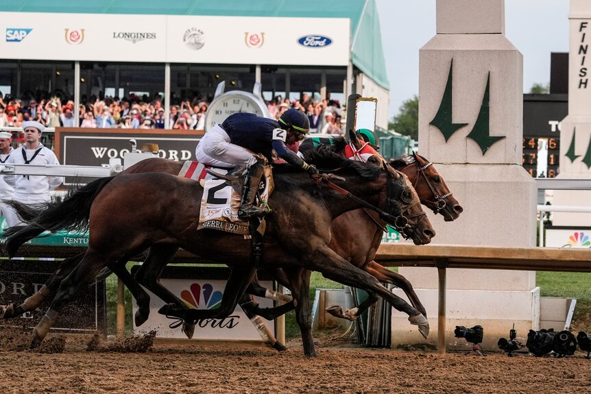 Three racehorses hit the finish line in a photo finish, with the horse furthest from picture winning.