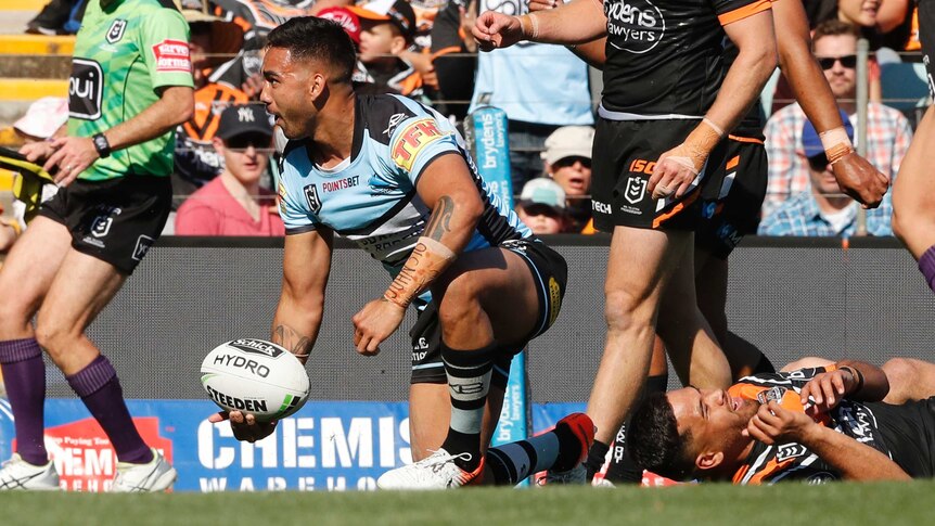 A male NRL player kneels as he holds the ball in his right hand after scoring a try.