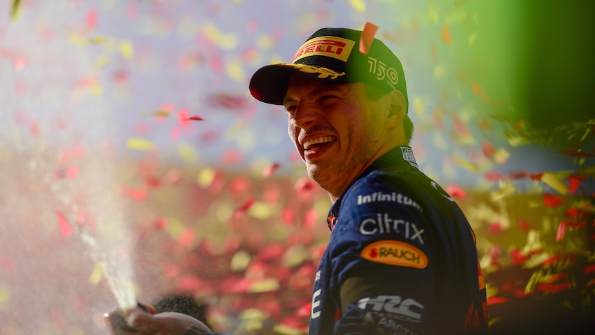 F1 driver Max Verstappen stands laughing as he pops a champagne cork and confetti falls around him.