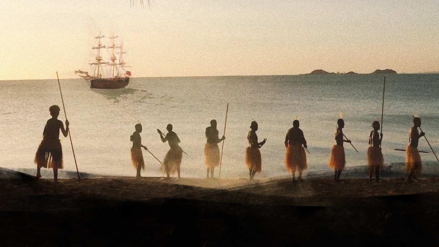 Aboriginal people line a coastline while HMB Endeavour floats in the distance close to shore.