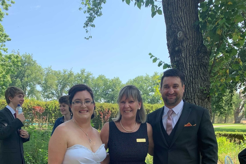 A smiling woman stands between a happy couple on their wedding day.