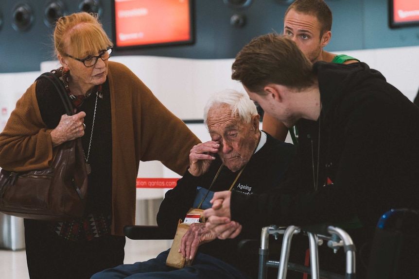 A man in a wheelchair surrounded by younger relatives in front of an airport check-in counter.