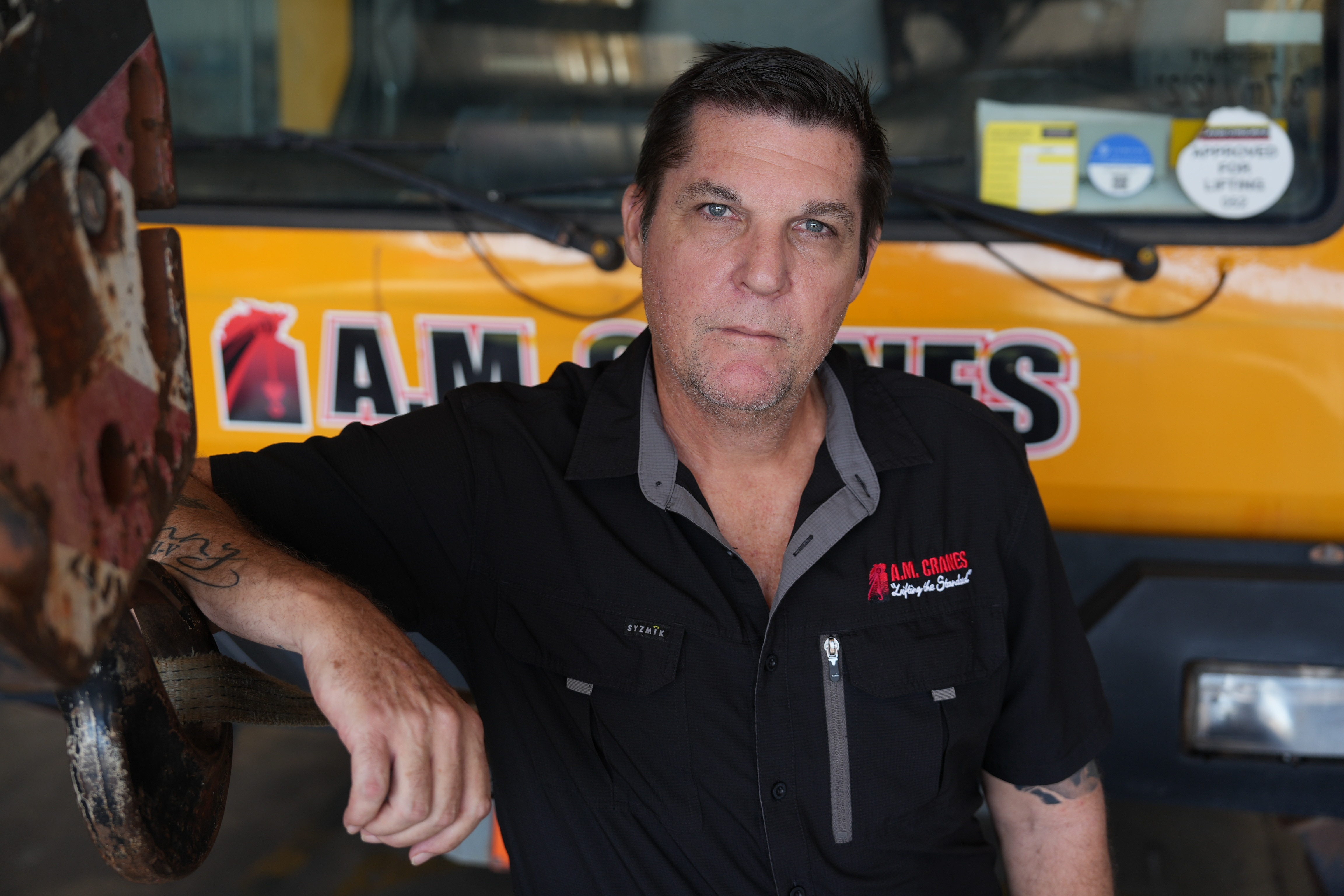 White man with black short cut hair, wearing a black polo shirt, stading in front of a yellow vehicle, with a serious expression