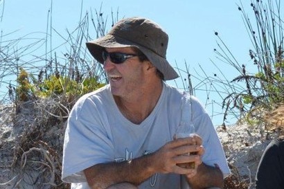 A man wearing a white t-shirt, hat and sunglasses sits in front of a sand dune laughing with a beer.