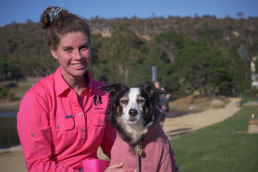 A young woman in a bright pink shirt holding and sitting next to a collie dog, smiling to camera.