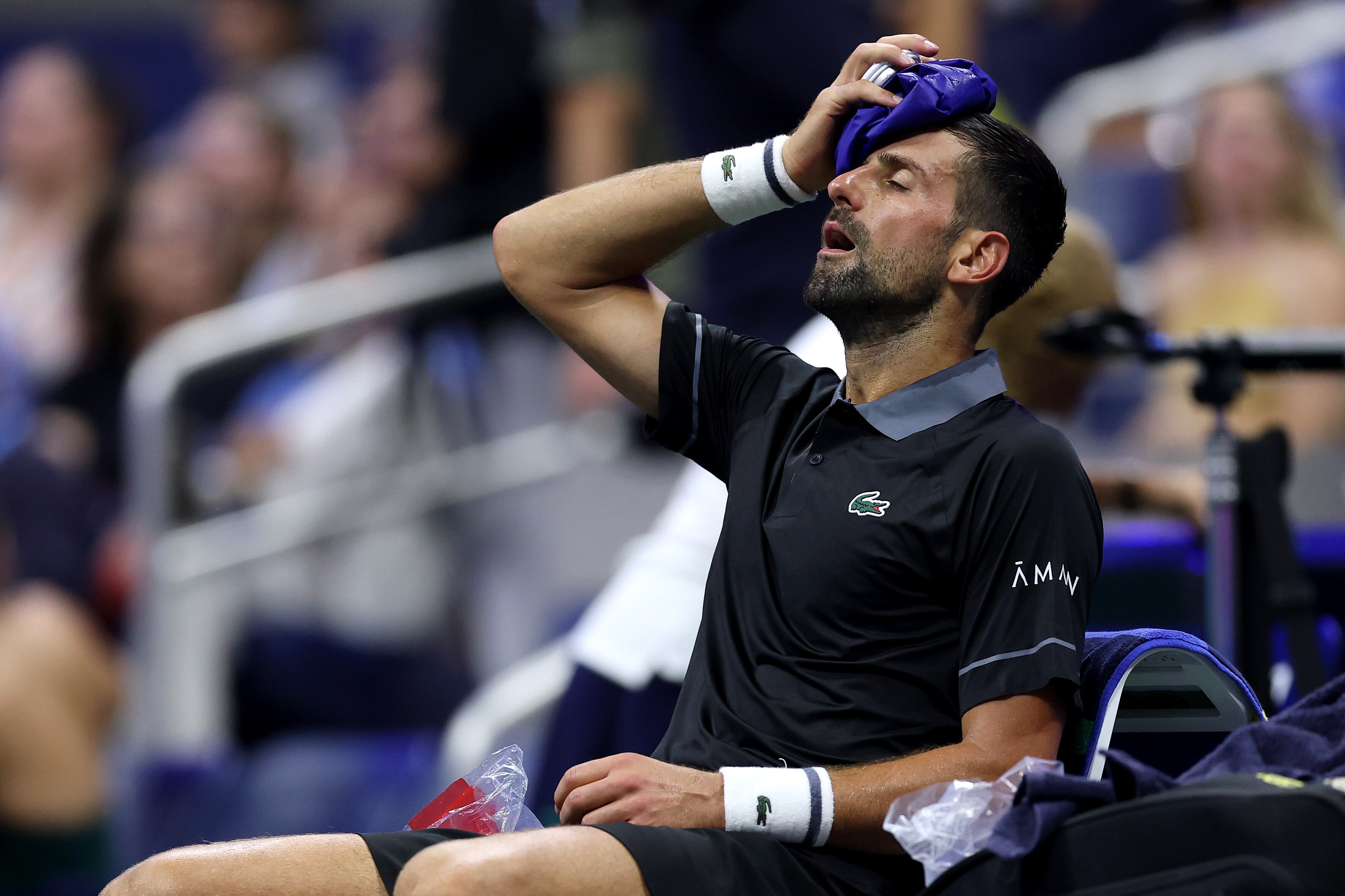 Novak Djokovic with an ice pack on his forehead and his eyes shut while sitting on the sidelines.