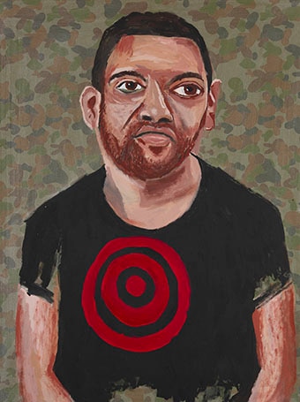 An Indigenous man sits against a camouflage background with a red target on his shirt.