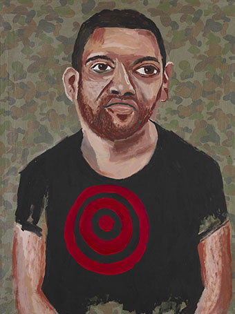 An Indigenous man sits against a camouflage background with a red target on his shirt.