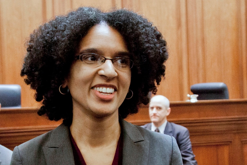 A black woman stands in a court room, smiling mid sentence.