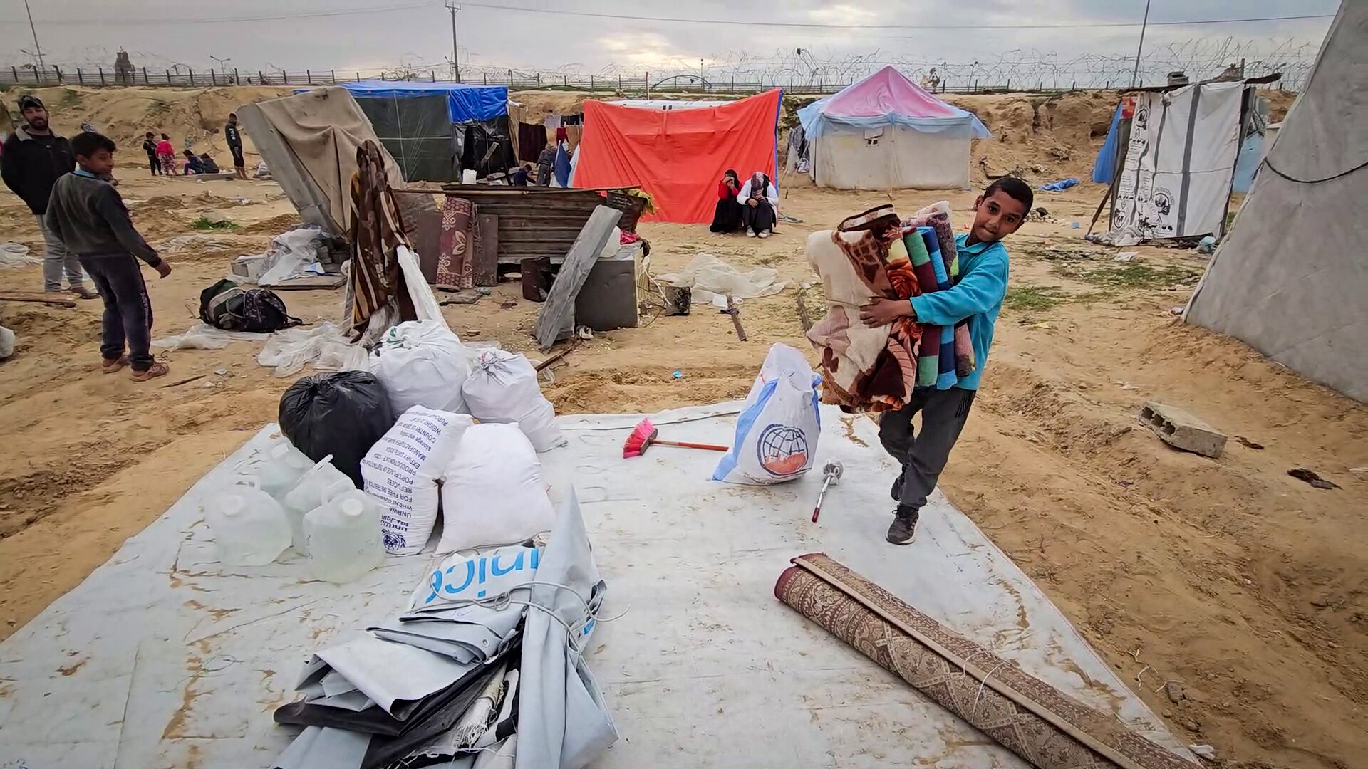 A young boy hugs a pile of blankets to his chest as he walks across a tarp with scattered belongings on it 