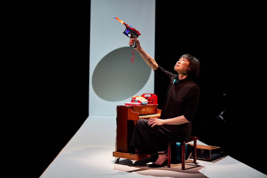 An East Asian woman sits on a chair in front of a tiny piano, with toys atop it. She holds a toy gun in the air.