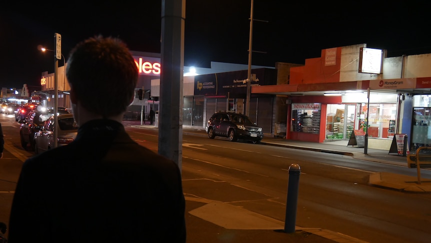 A man walks down a suburban shopping strip at night.