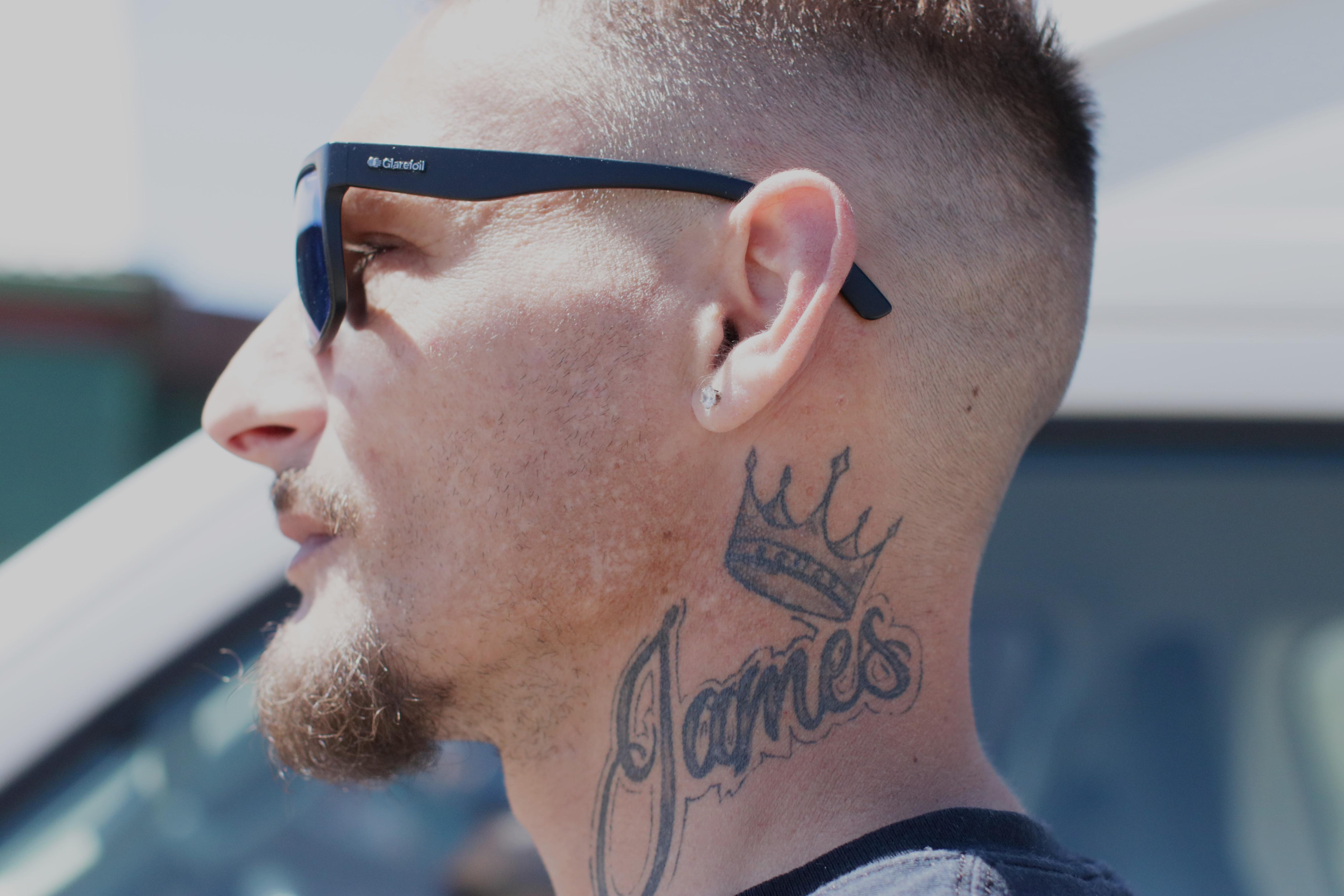 Close up side profile of man with goatie, moustache, sunglasses, tattoo on neck of crown and word James
