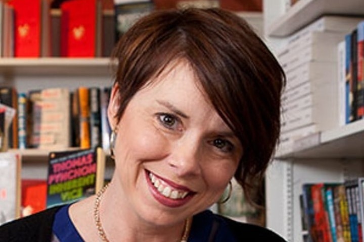 Woman with short brown hair smiles while sitting in front of book case. 