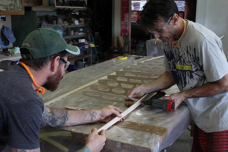 Two men using a nail gun to attach wooden ribs to a wooden frame.