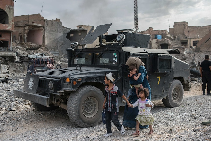 Women and children walk past a special forces vehicle.