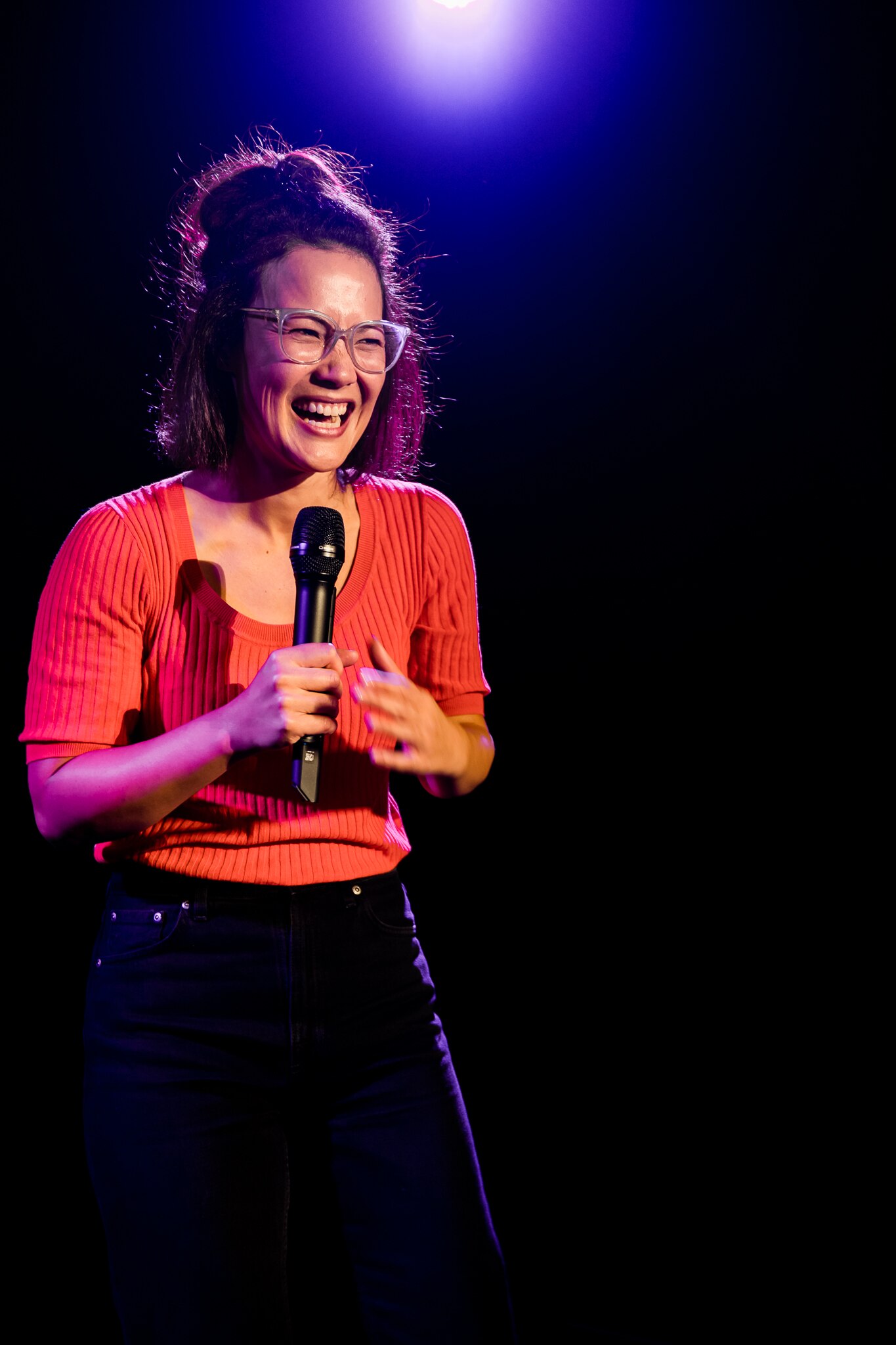 A smiling 30-something Asian Australian woman is on stage in a dark theatre. She's wearing glasses and holding a microphone.