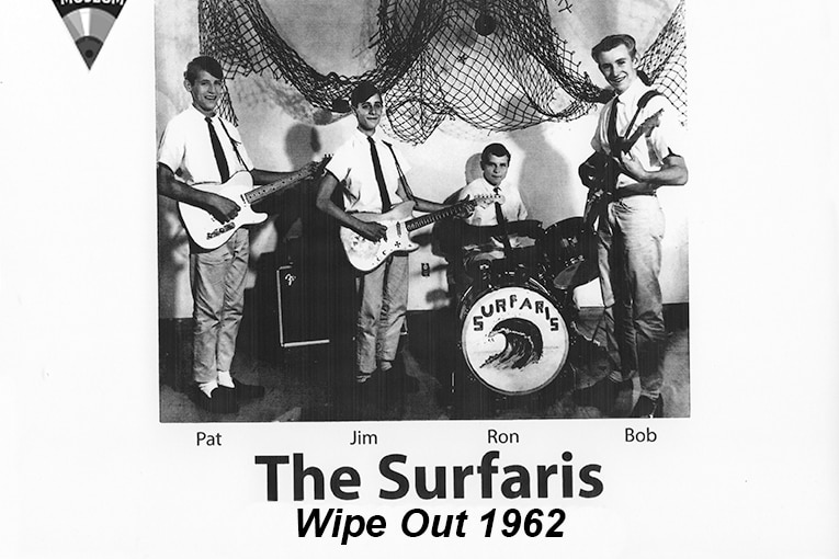 A group of four young men in shirts and ties, three holding guitars and one at a drum kit with the word 'Surfaris' on the front