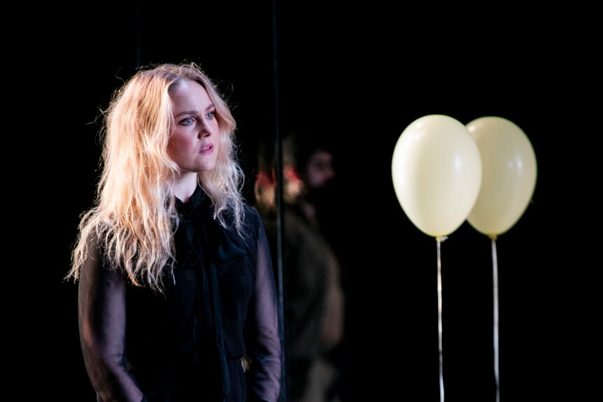 White woman with long blonde hair stands onstage with hands clasped in black dress and boots; a white helium balloon behind her.