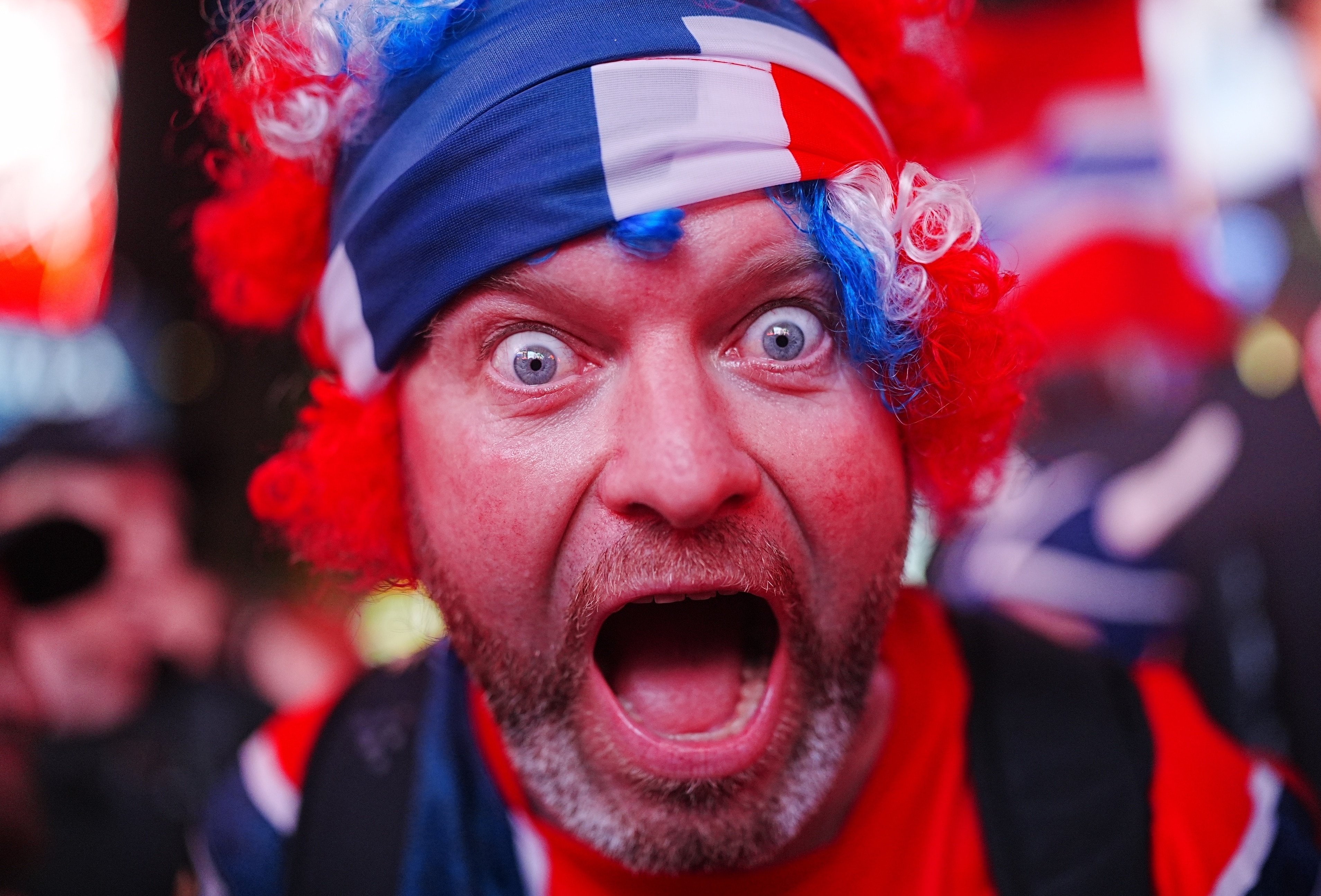 A man with a red wig and headband screams with joy in an extreme close up