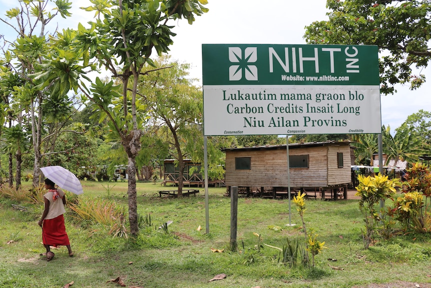 A woman with an umbrella walks past a sign for the company NIHT in a PNG village.