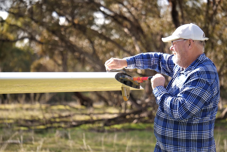 A man in a white cap and blue checked shirt adjusts a plane wing.