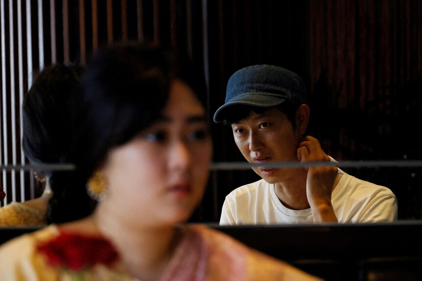 Un jeune japonais se reflète dans un miroir tout en parlant à une jeune japonaise en sari. 