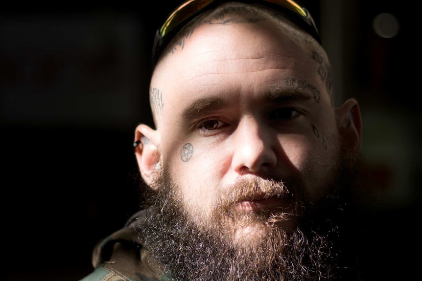 A bearded man with face tattoos look forward with a pensive expression.
