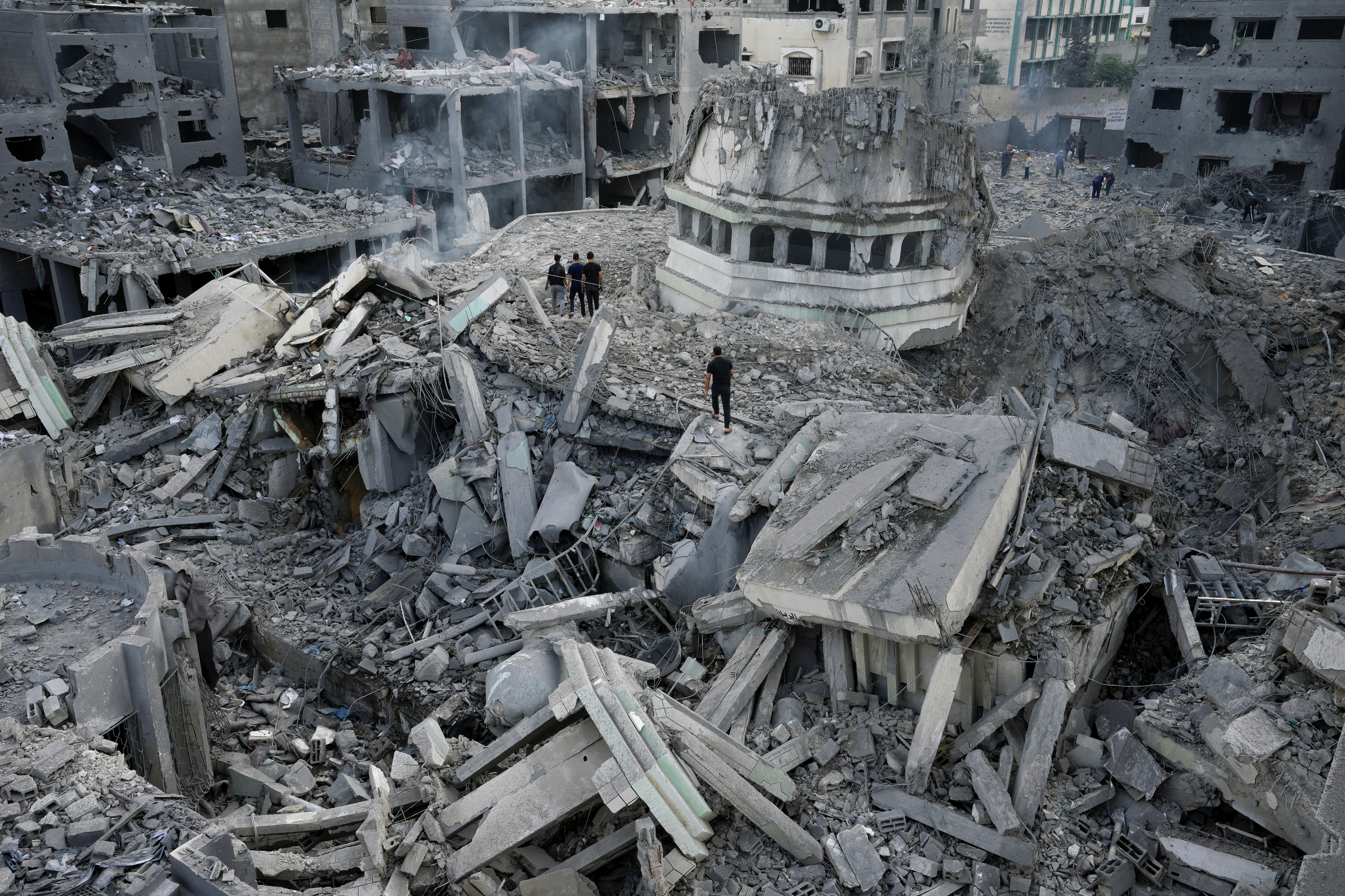 Four people in black stand surrounded by vast piles of grey rubble of destroyed buildings.