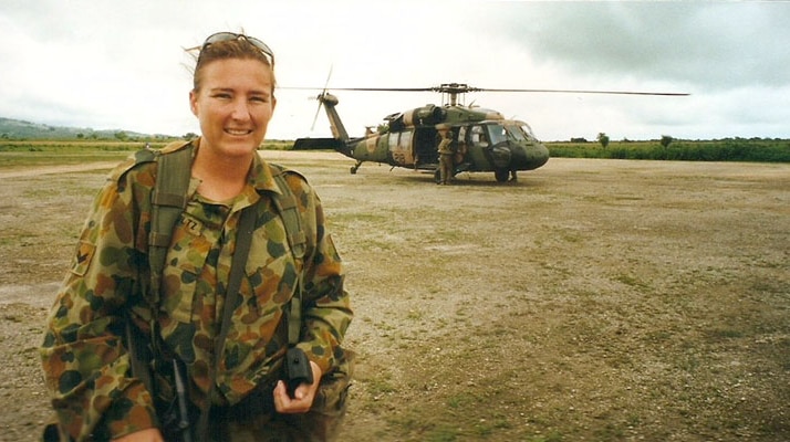 A woman in army camouflage stands on a tarmac with an army helicopter and empty fields in the background.