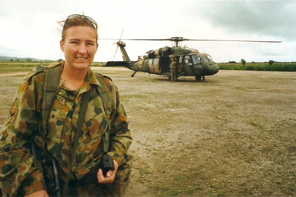 A woman in army camouflage stands on a tarmac with an army helicopter and empty fields in the background.