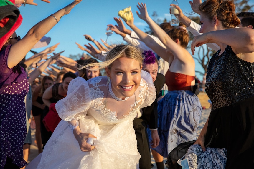 A woman in a wedding dress with a glowing smile runs through a crowd at the beach.