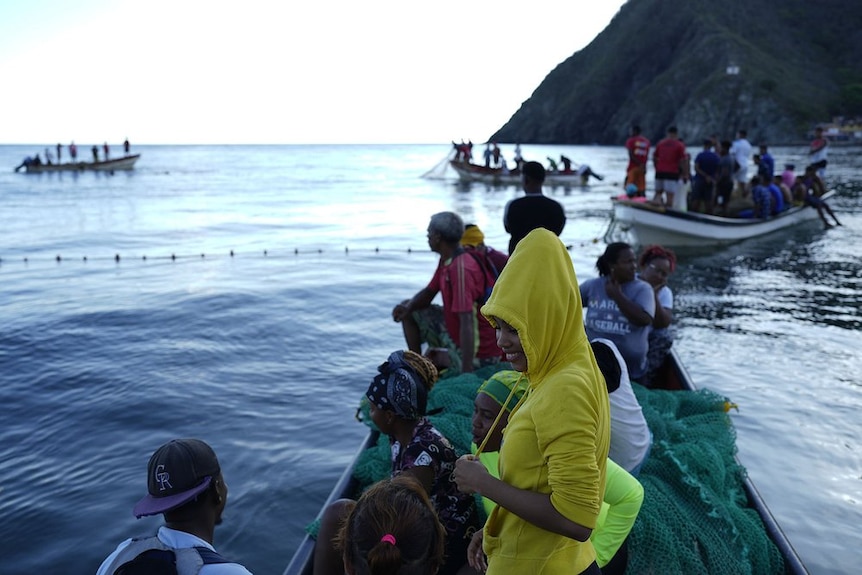 A woman wearing a hoodie stands in a small fishing boat filled with people and fishing nets, other boats are in the background