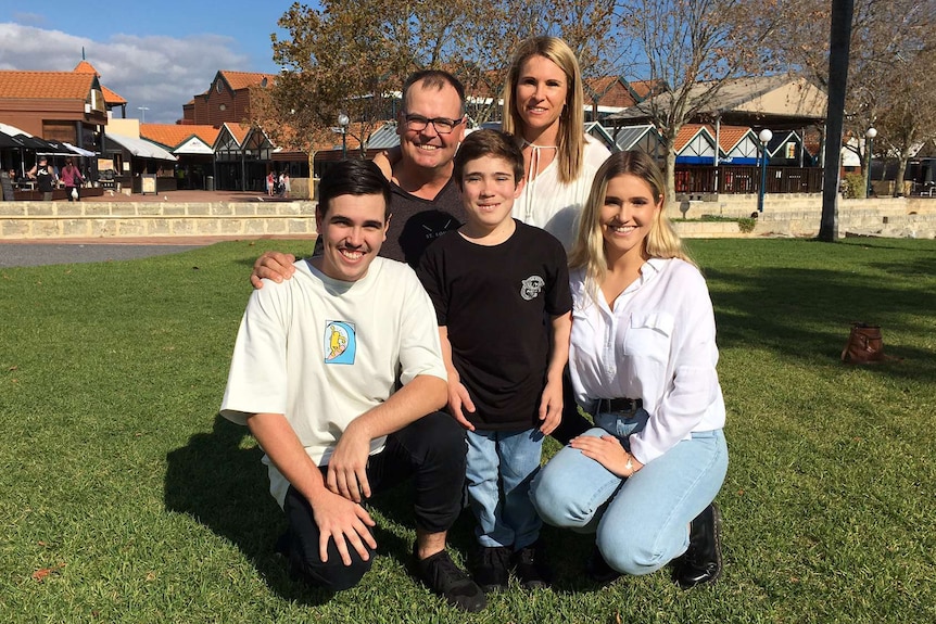 Coby Antonio stands wearing a black t-shirt and jeans surrounded by his crouching family on both sides.