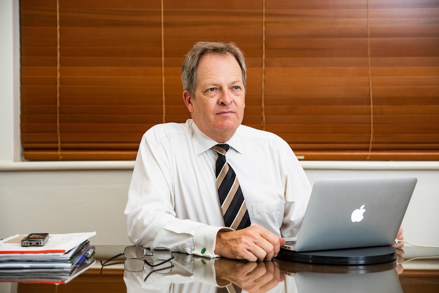 A man in a white shirt with a black and gold tie looks to the side while sitting at a desk with a glass top.