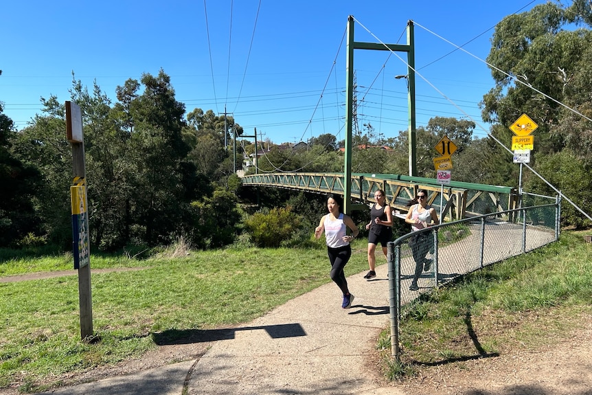 Three women run close to each other down a nature trail, a green footbridge is in the background.