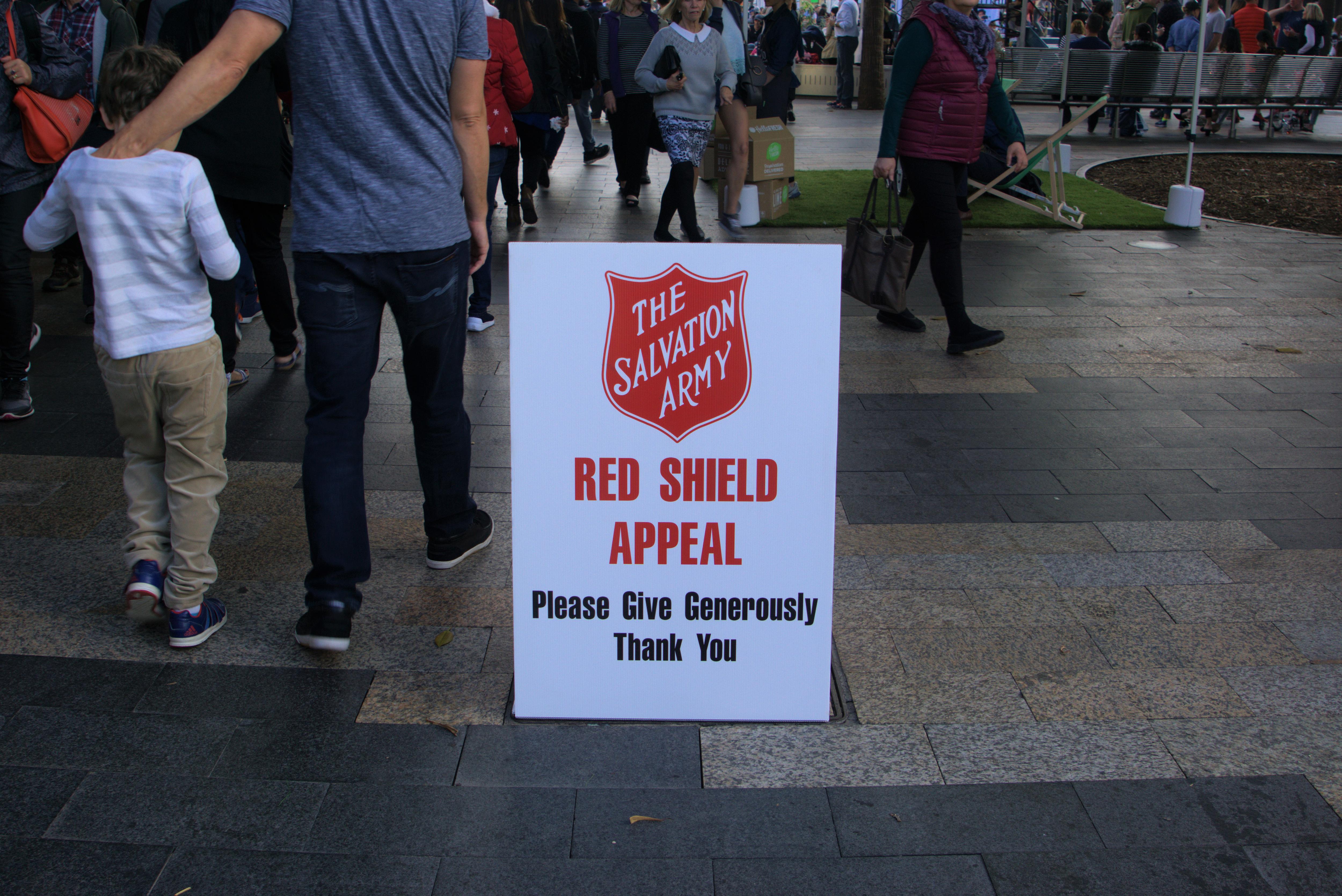 People walk past a white sign with red writing for the Red Shield Appeal by Salvation Army.