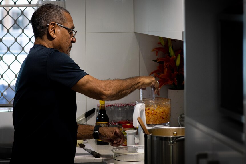 A man stands in a kitchen and is mixing chillies, ginger and garlic in a food processer. 