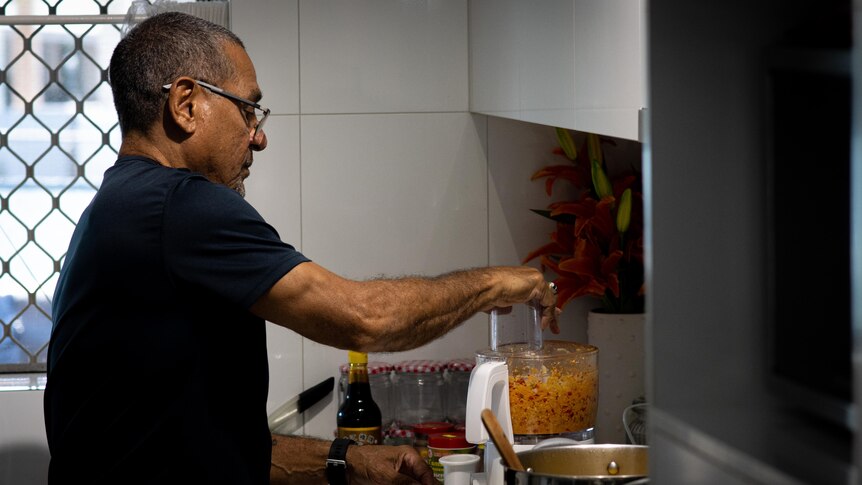 A man stands in a kitchen and is mixing chillies, ginger and garlic in a food processer. 