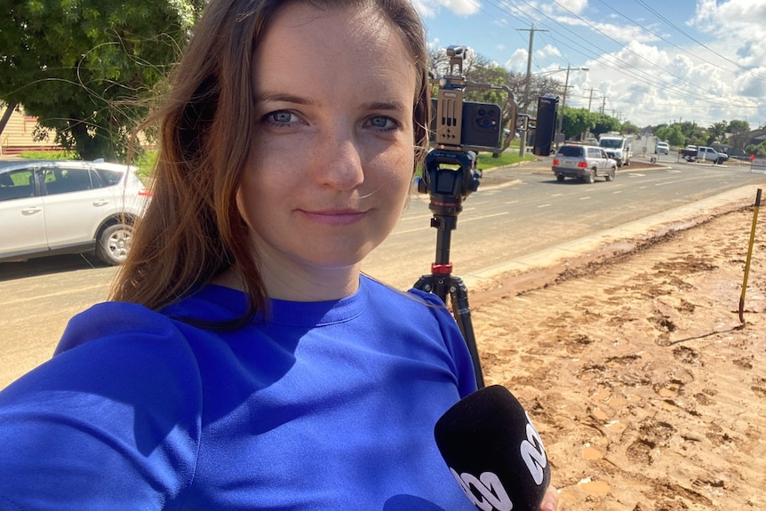 Woman holding a microphone standing in front of a mobile camera kit on a tripod on a street.