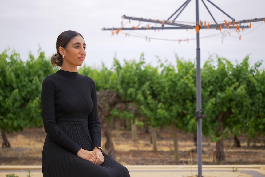 A woman wearing a black dress sits in front of grape vines and a clothes line