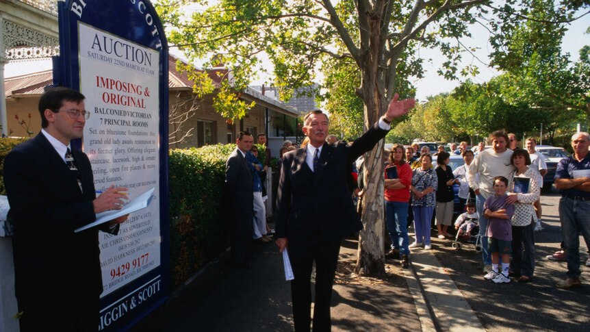 A crowd of people gather on the street while an auctioneer takes bids in Melbourne. The street is lined with leafy trees