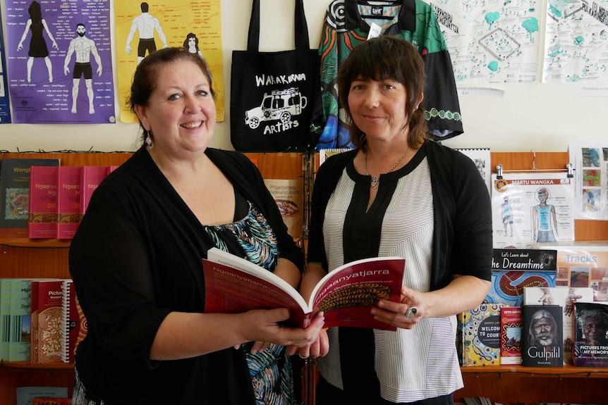 Two women hold a book together and stand in front of  book shelves and a wall of educational charts.