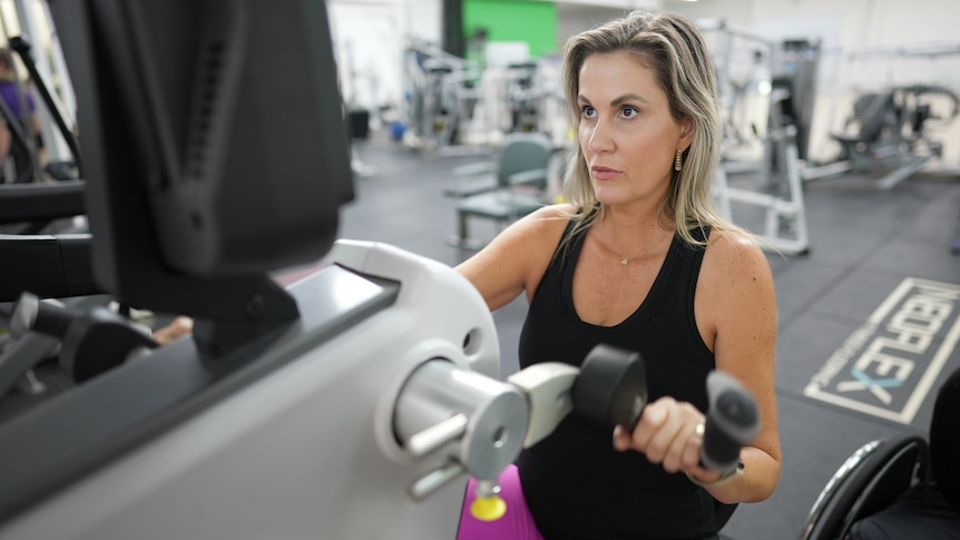 Karni Liddell, dressed in black and purple workout gear, holds the handles of a piece of exercise equipment while seated.