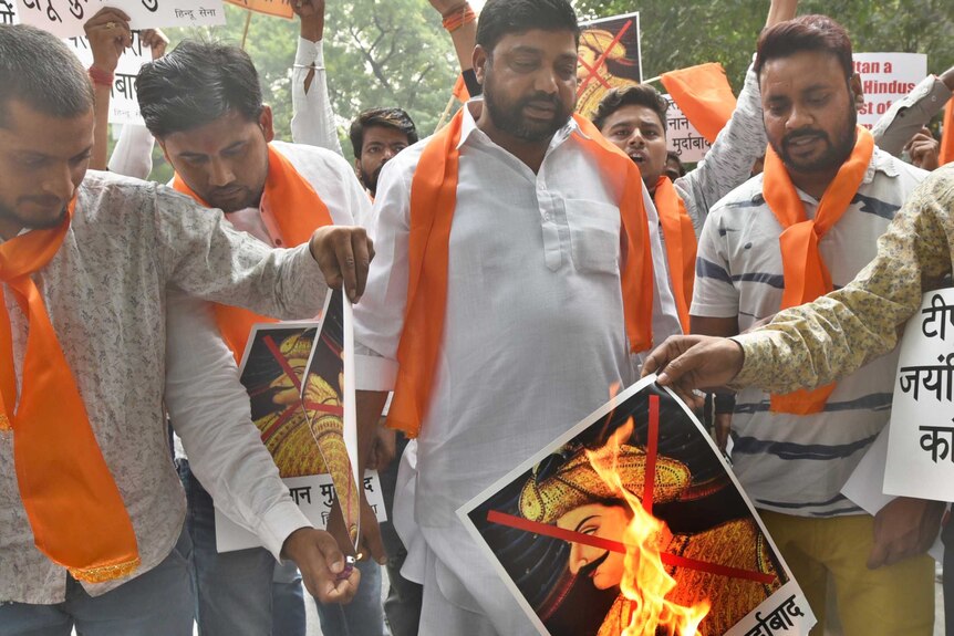 Men with orang scarves hold signs and set fire to a picture of Tipu Sultan with a red cross on it.