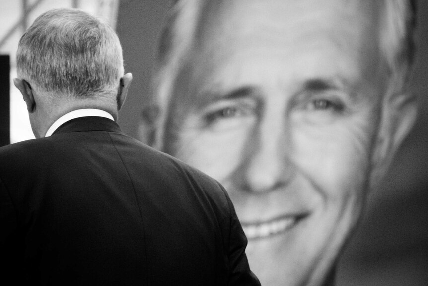 Prime Minister Malcolm Turnbull turns towards a large sign of his smiling face.